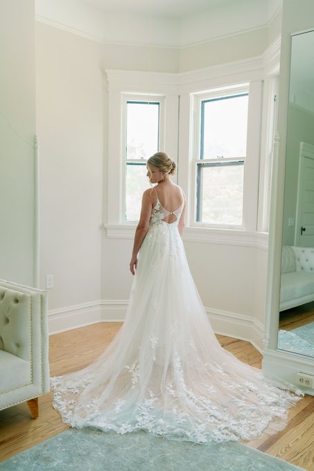 Bride standing at a bay window with her lace train spread behind her
