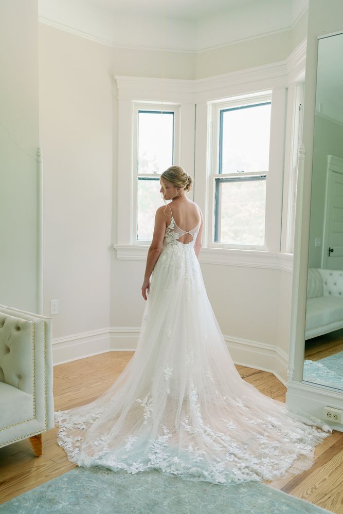 Bride standing at a bay window with her lace train spread behind her