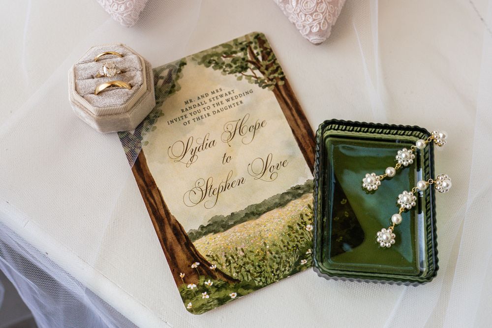 Invitation suite in green and gold beside the wedding rings and earrings