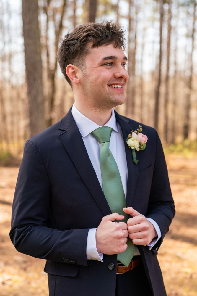 The groom in a navy suit and sage tie fastening his jacket